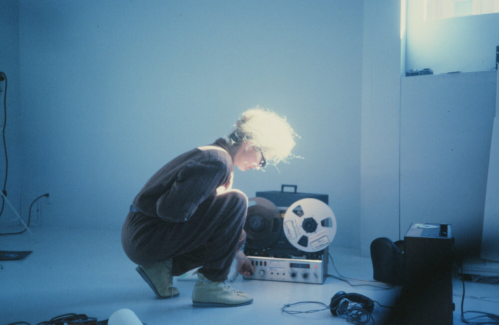 In einem hellblauen Raum hockt eine hellhaarige Frau im Overall vor einem Gerät mit Filmrolle.